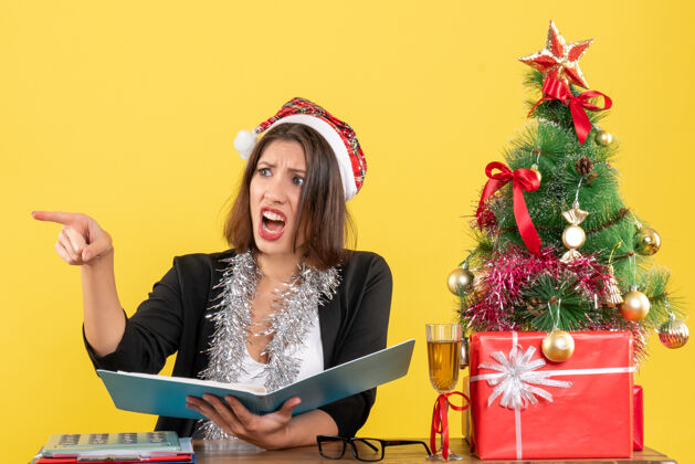 文件穿着西装 戴着圣诞老人帽和新年装饰品的商务女士正在检查文件 指着什么东西 坐在办公室的桌子旁 桌子上放着圣诞树帽子桌子笔记本电脑