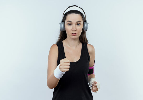 健身戴着耳机 戴着智能手机袖标的年轻健身女士站在白色背景下 严肃地看着摄像机面部年轻耳机