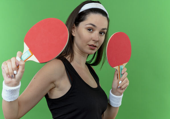 相机戴着头巾拿着球拍打乒乓球的年轻健身女士站在绿色背景下 自信地看着镜头网球球拍年轻