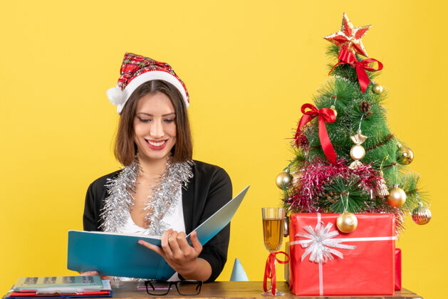漂亮穿着西装 戴着圣诞老人帽和新年装饰品的商务女士正在阅读文件 坐在办公室的桌子旁 桌上放着圣诞树圣诞老人帽子树