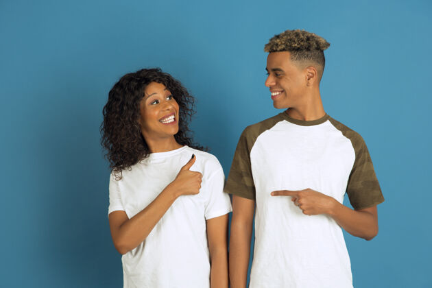 朋友年轻感性的非裔美国人 穿着白色休闲服 在蓝色背景上摆姿势美丽的情侣人类情感的概念 面部表情 关系 互相指指点点的广告衣服男性情侣