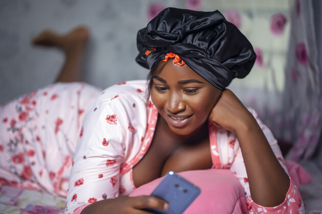 非洲一位年轻的非洲裔美国女性戴着缎子帽子在床上用手机的画像美国圆形浴衣