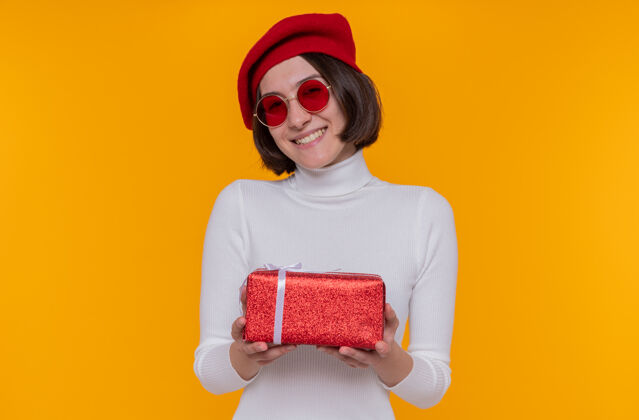 高领毛衣一个留着短发 头戴贝雷帽 头戴红太阳镜 头戴白高领毛衣的年轻女子 手里拿着一份快乐而积极的礼物穿女士橙色