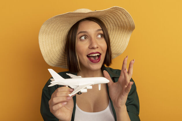 飞机一个戴着沙滩帽的兴奋的漂亮女人拿着飞机模型 看着橙色墙上孤立的一面举行女人帽子