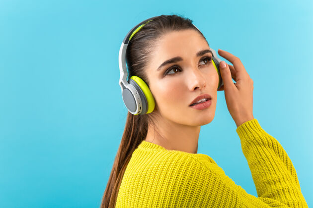 服装迷人时尚的年轻女子戴着无线耳机听音乐快乐地穿着黄色针织毛衣五颜六色的时尚造型隔离在蓝色的墙上休闲欢乐耳机