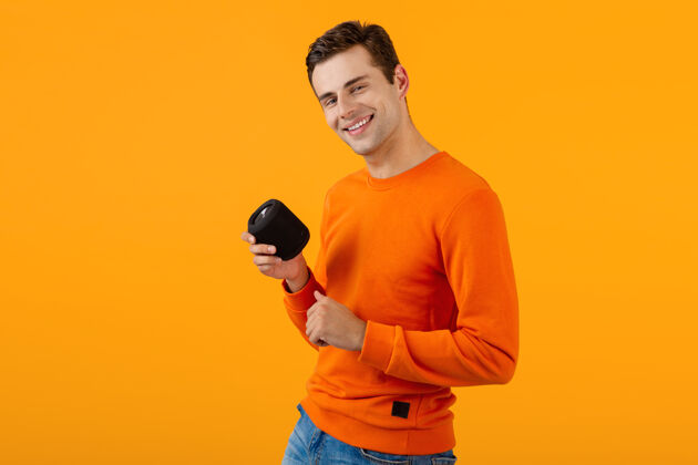 姿势时尚微笑的年轻人穿着橙色毛衣手持无线扬声器快乐地听音乐娱乐扬声器有趣