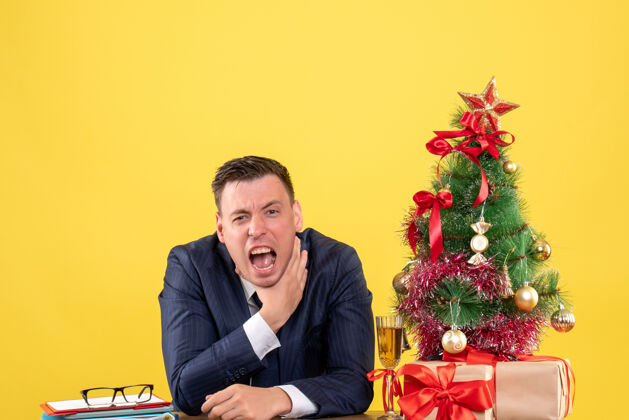 男人前视图愤怒的男人用手勒死自己坐在圣诞树和黄色背景上的礼物旁边的桌子上树生意人礼物
