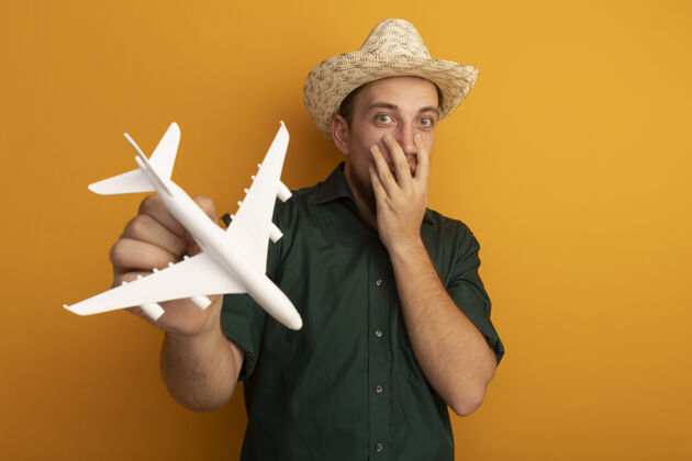 帽子戴着沙滩帽的金发帅哥惊讶地把手放在脸上 把飞机模型孤立地放在橙色的墙上飞机市民模特