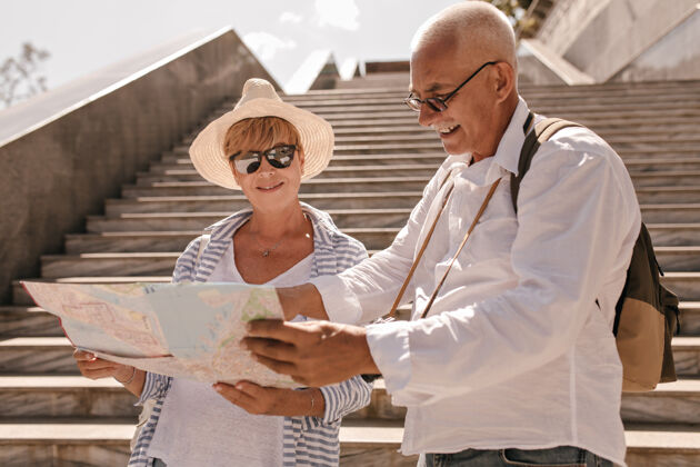 游客戴着眼镜 穿着浅色衬衫 背着背包的灰头发男人和戴着帽子 穿着蓝色条纹衣服的现代女人一起看地图聪明祖父母朋友
