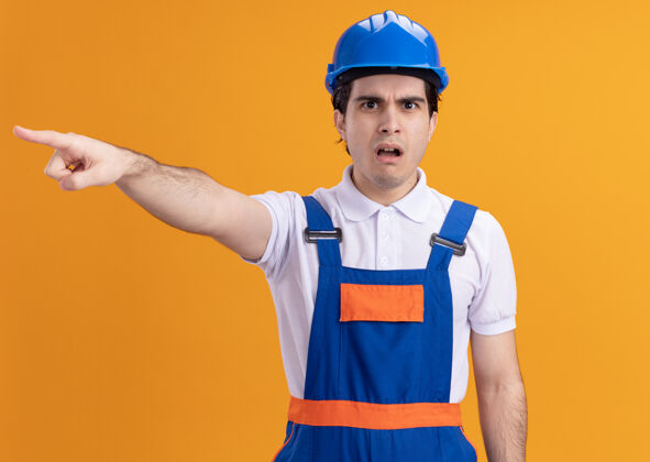 建筑年轻的建筑工人穿着建筑制服 戴着安全帽 困惑地看着前面 担心地用食指指着橘色墙上的东西目录安全制服