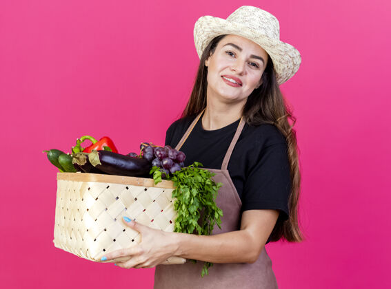 蔬菜微笑美丽的园丁女孩穿着制服 戴着园艺帽 拿着蔬菜篮子 在粉红色的背景上隔离开来穿微笑女孩