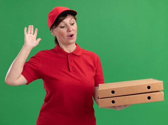 年龄身穿红色制服 头戴鸭舌帽的中年送货妇女举着披萨盒 高举手臂站在绿色的墙上 高兴而兴奋地看着披萨盒中间快乐披萨