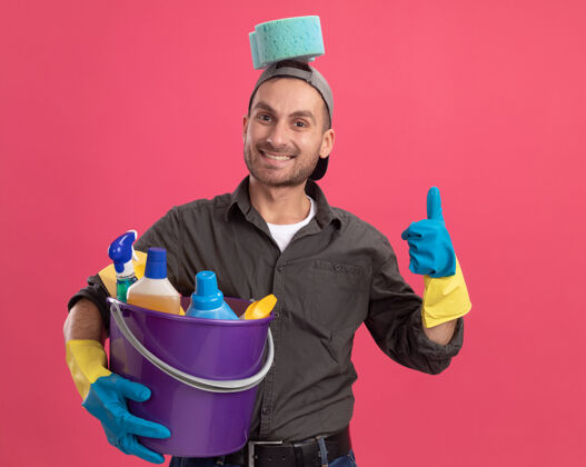 男人快乐的年轻人穿着休闲服 戴着橡胶手套 戴着帽子 手里拿着带清洁工具的水桶 头上戴着海绵 微笑着竖起大拇指站在粉色的墙上穿着微笑表演