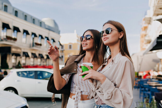 友谊戴着太阳眼镜的漂亮女孩们拿着咖啡 用智能手机 站在户外微笑着女人女士情感