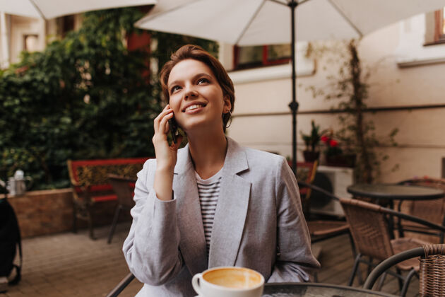年轻快乐的女士在咖啡馆休息 讲电话迷人的短发女子穿着灰色夹克 微笑着 外面冷咖啡馆咖啡馆女人