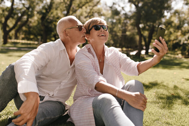 公园一个活泼酷的女人 一头金发 穿着现代条纹衬衫 戴着眼镜 坐在草地上 微笑着和公园里的白发男人自拍电话夫妇老年人