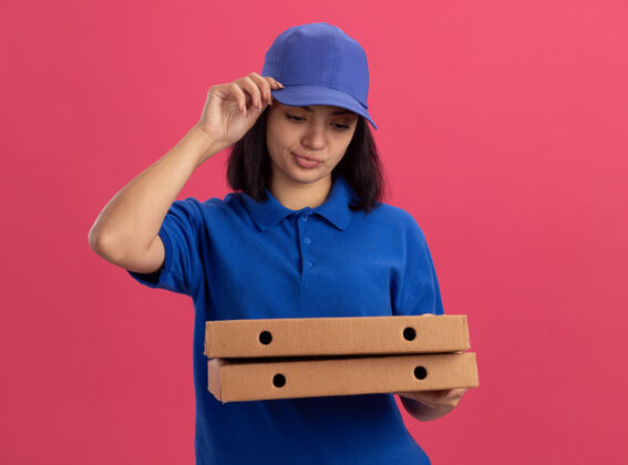 站着身穿蓝色制服 头戴鸭舌帽 拿着披萨盒的年轻送货员站在粉红色的墙上 歪歪扭扭地说着失望的表情女孩制服年轻