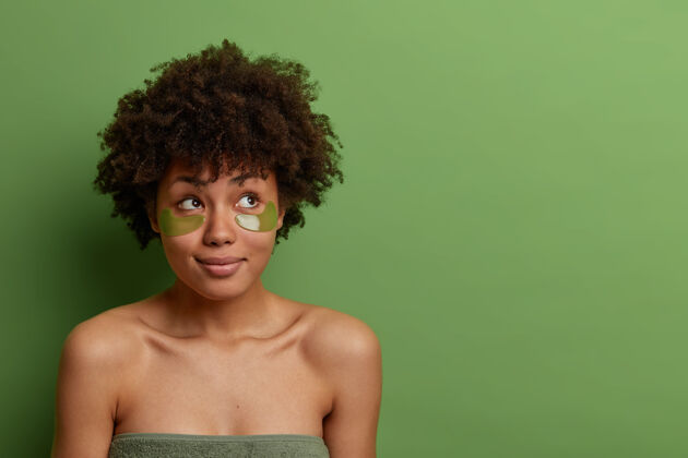 肩膀美丽健康的生活方式理念体贴美丽的深色皮肤的女人在眼睛下敷上眼罩 满意有效的新化妆工具裹在毛巾里 看着隔离在绿色墙上的一边女人思想思考