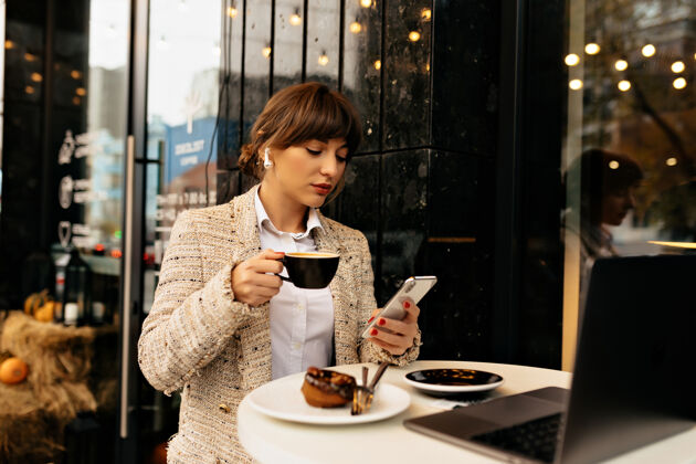 集中穿着夹克的年轻时尚商务女性 使用智能手机和笔记本电脑 听音乐和喝咖啡社会工作微笑