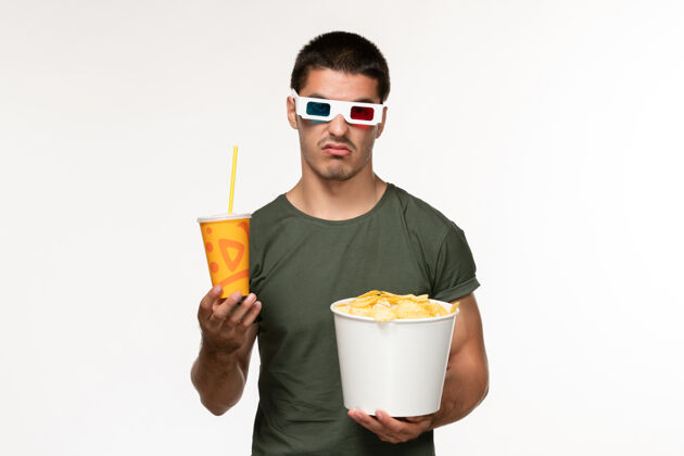 电影正面图身穿绿色t恤的年轻男性手持马铃薯cips苏打水 戴着d墨镜 在白色墙上拍摄电影《男性孤独》电影院电影肖像视图