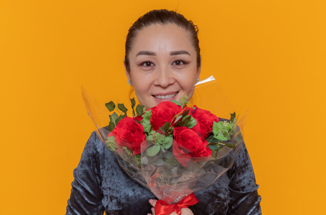 玫瑰快乐的亚洲女人捧着一束红玫瑰望着前方微笑着站在橙色的墙上欢庆国际妇女节庆祝举办微笑