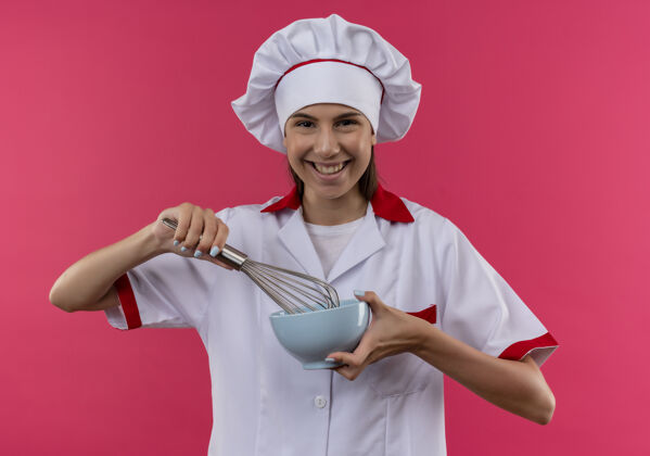 复制身着厨师制服的年轻微笑的白人厨师女孩拿着打蛋器和碗放在粉红色的地板上 留有复印空间搅拌制服粉红