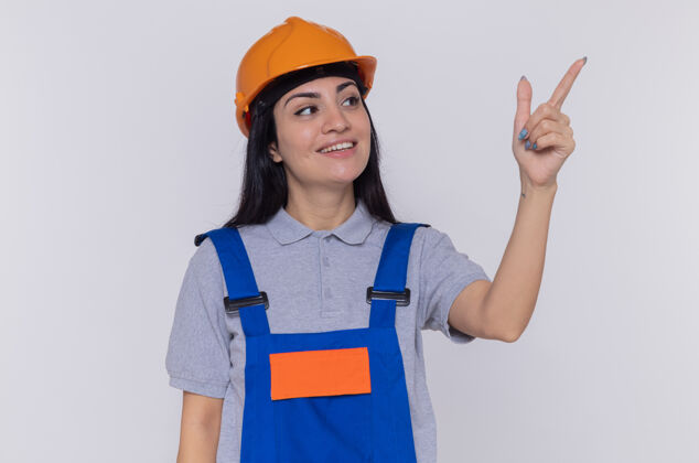 制服身穿建筑制服 头戴安全帽的年轻建筑工人面带微笑 用食指指着站在白墙上的东西女人安全旁边