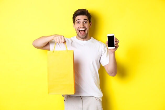 表情手机银行和现金返还的概念年轻快乐的家伙拿着购物袋 展示智能手机屏幕 黄色背景男人黄色自信