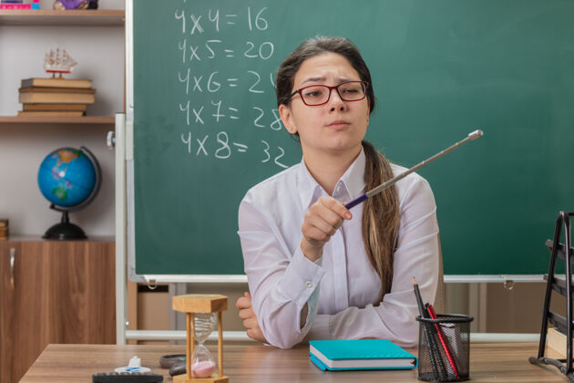 解释戴着眼镜的年轻女老师面带严肃的表情 指着坐在教室黑板前的课桌上讲解课文的东西穿学校前面