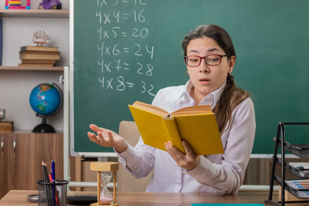 眼镜戴眼镜的年轻女教师拿着备课书 困惑地坐在教室黑板前的课桌上困惑黑板拿着