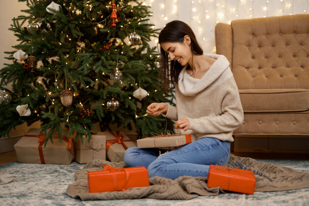 年轻可爱的女人打开圣诞礼盒 坐在家里圣诞树旁的地板上天哪花环坐着