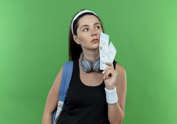 立场戴着头巾 背着背包 拿着机票 表情严肃地站在绿色墙壁上的年轻健身女士背包健身女人