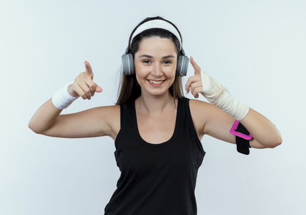 年轻人戴着头戴耳机和智能手机袖标的年轻健身女士微笑着 用食指指着站在白墙上的一边手机目录壁板