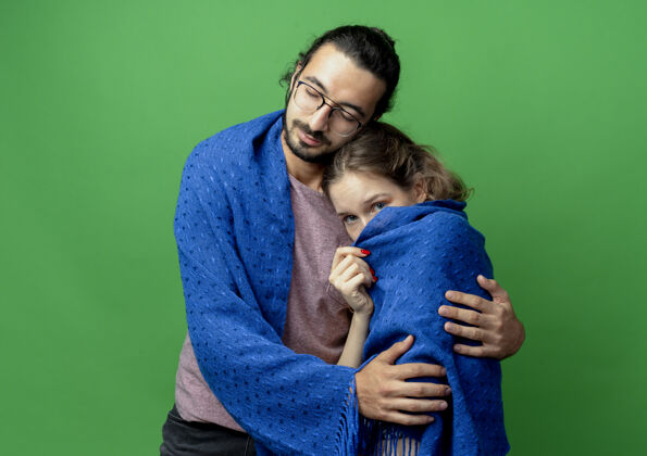 年轻一对年轻的男女 幸福的男人拥抱着他心爱的女友 把她裹在温暖的毯子里 站在绿色的墙上拥抱毯子温暖