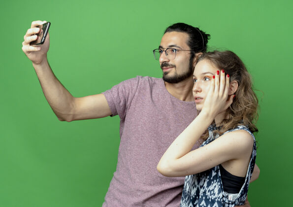 照片一对年轻的男女 快乐的男人用他的智能手机拍下他们站在绿色的墙上男人情侣手机
