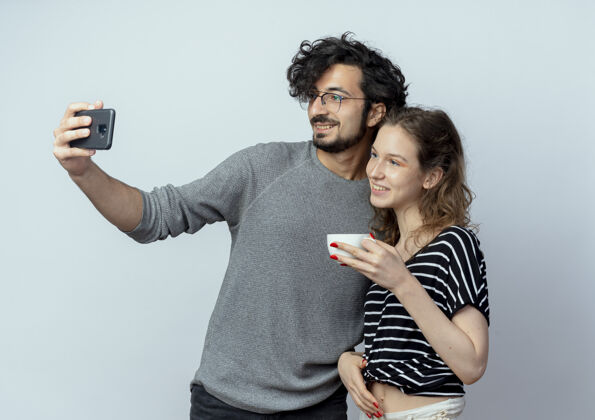 女人一对年轻的男女 快乐的男人用智能手机拍下了他们的照片 而他的女朋友站在他旁边 隔着白墙喝着咖啡拿着站着手机