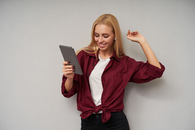 个人电脑乐观的年轻迷人的女士 留着一头长发 在平板电脑上听音乐 举手愉快地跳舞 身穿紫红色衬衫和浅灰色背景的白色t恤年轻漂亮爱情