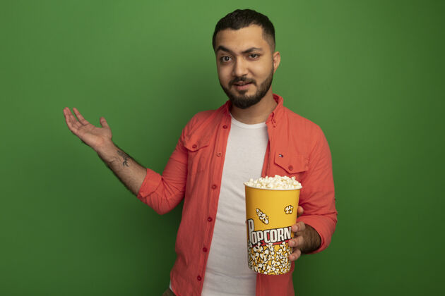 水桶一个留着胡子的年轻人穿着橘色衬衫 手里拿着装满爆米花的桶 胳膊站在绿色的墙上爆米花站着胡子