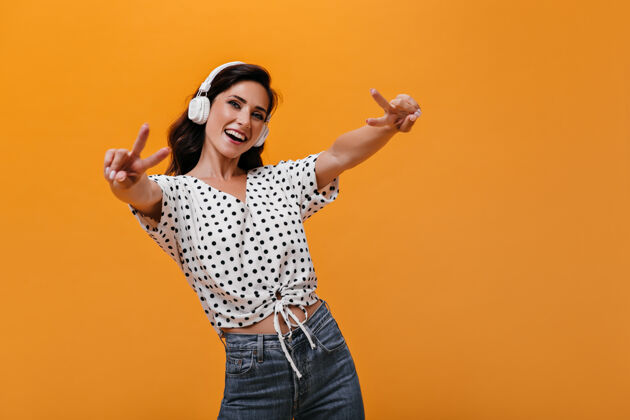 女性戴着耳机的女孩表现出和平的迹象 戴着耳机听音乐微笑的女人 深色波浪发 穿着白色圆点衬衫 玩得很开心橙色背景人音乐