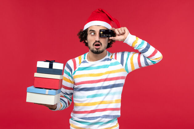 漂亮正面图年轻男子手持礼物和银行卡在红墙上压岁钱红表演圣诞节举行