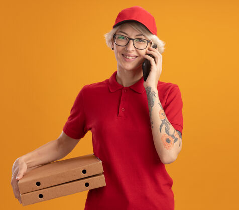 手机身穿红色制服 戴着帽子 戴着眼镜 拿着披萨盒 拿着手机 站在橘色的墙上开心地微笑着的年轻送货员电话制服站着