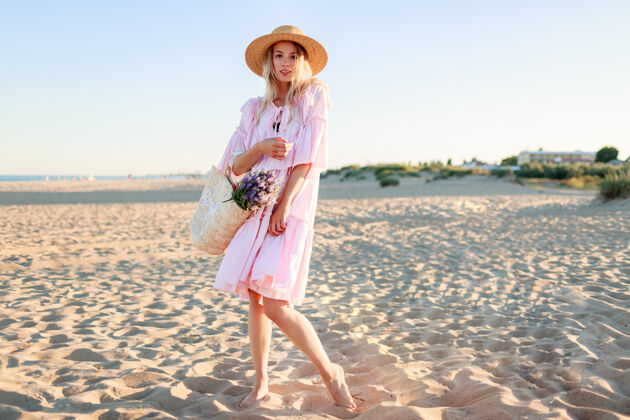 海滩金发女孩穿着可爱的粉色裙子在沙滩上跳舞 手里拿着草包和鲜花美丽优雅风