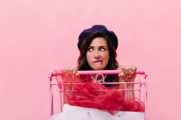 购物车戴贝雷帽的女孩的肖像 她喜欢购物和手推车戴贝雷帽的黑色卷发女人在粉色背景下摆姿势购物情绪乐趣