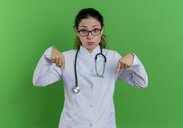 指向年轻的女医生穿着医用长袍 戴着听诊器 戴着眼镜 孤独地指着绿色的墙上看医生长袍