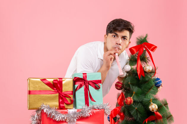 微笑前视图年轻人周围的圣诞礼物和节日树上的粉红色背景圣诞树粉色风景