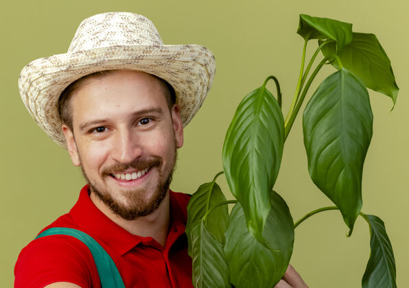 植物特写镜头中的快乐年轻英俊的斯拉夫园丁制服和帽子举行植物隔离橄榄绿墙看特写斯拉夫