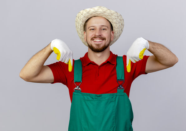 花园微笑的年轻英俊的斯拉夫园丁穿着制服戴着园艺手套和帽子指着隔离在白色墙上微笑制服指尖