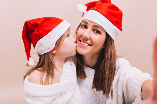 女人可爱的小女孩亲吻着戴着类似圣诞老人帽子的姐姐 女人在孤零零的墙上和女儿自拍 真是感人至深年轻小惊喜