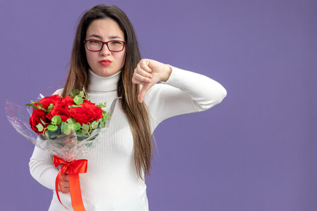 情人节穿着休闲服的年轻漂亮女人手持一束红玫瑰看着相机不高兴地竖起大拇指站在紫色的墙上庆祝情人节看拇指花束