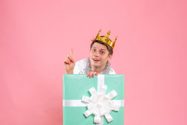 年轻男性正面图：年轻男性在粉色背景上戴着皇冠礼物丝带盒子